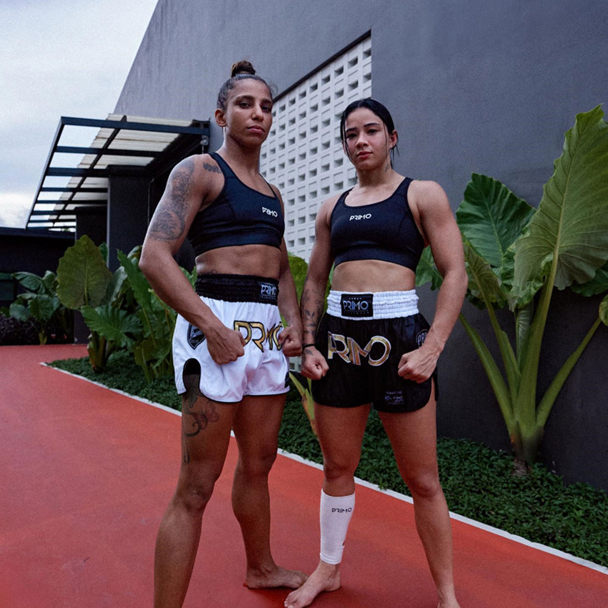 Two women in athletic wear standing on a red surface with greenery and a building in the background.